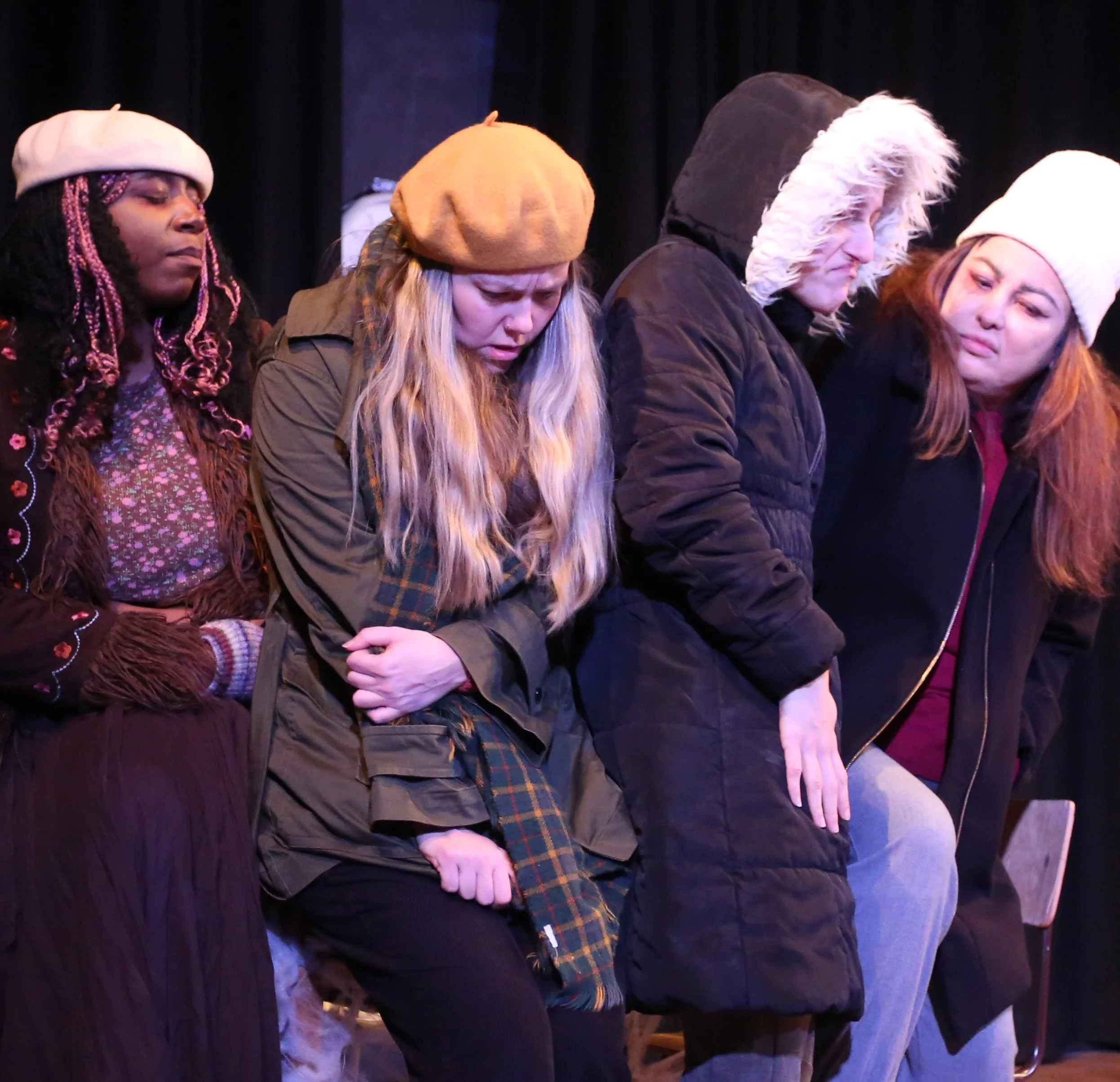 Four woman sit on the edge of a table huddled together. They look unhappy and cold, they wear many layers. The lighting is icy.