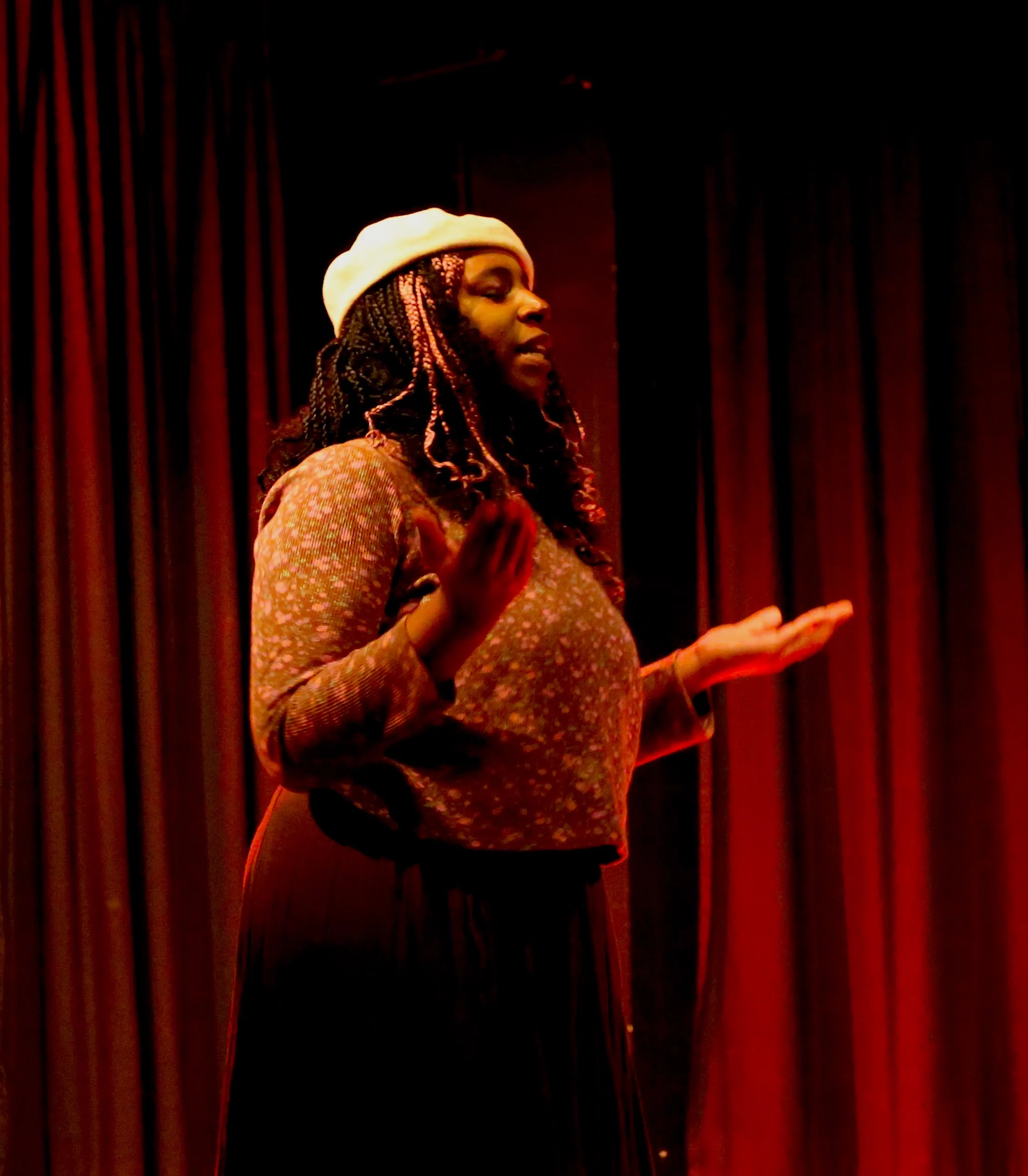 A woman stands on stage with her hands out and palms up. Her eyes are closed. She is wearing a white beret and brown loose clothing. The lighting washes her and the background in red.