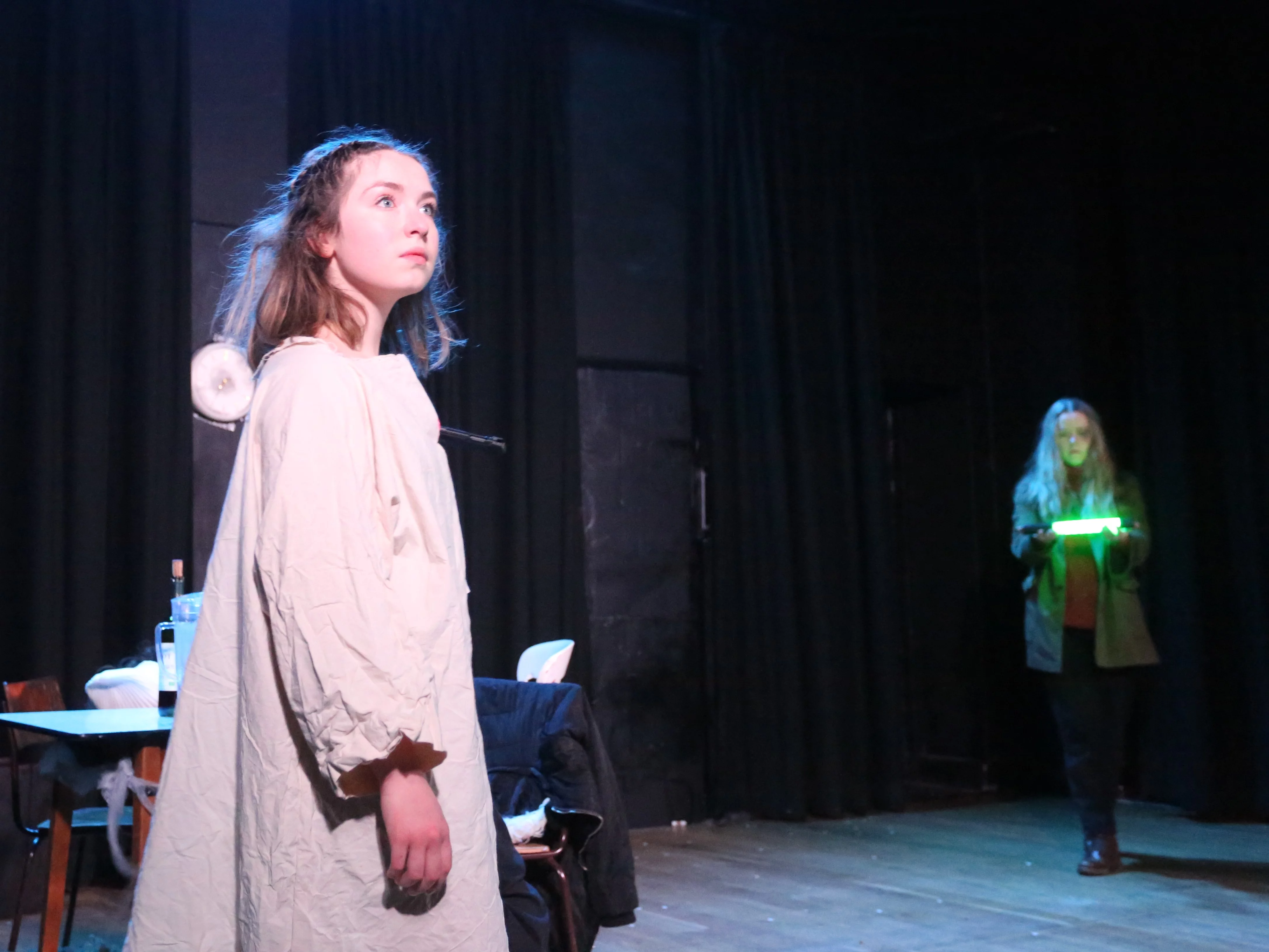 A woman stands alone onstage, she wears a white shroud. There is a messy table behind her. The slighting is white and stark.