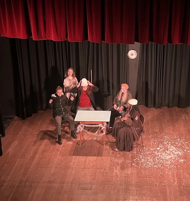A stage photo of four women around a table, with another woman walking around them.