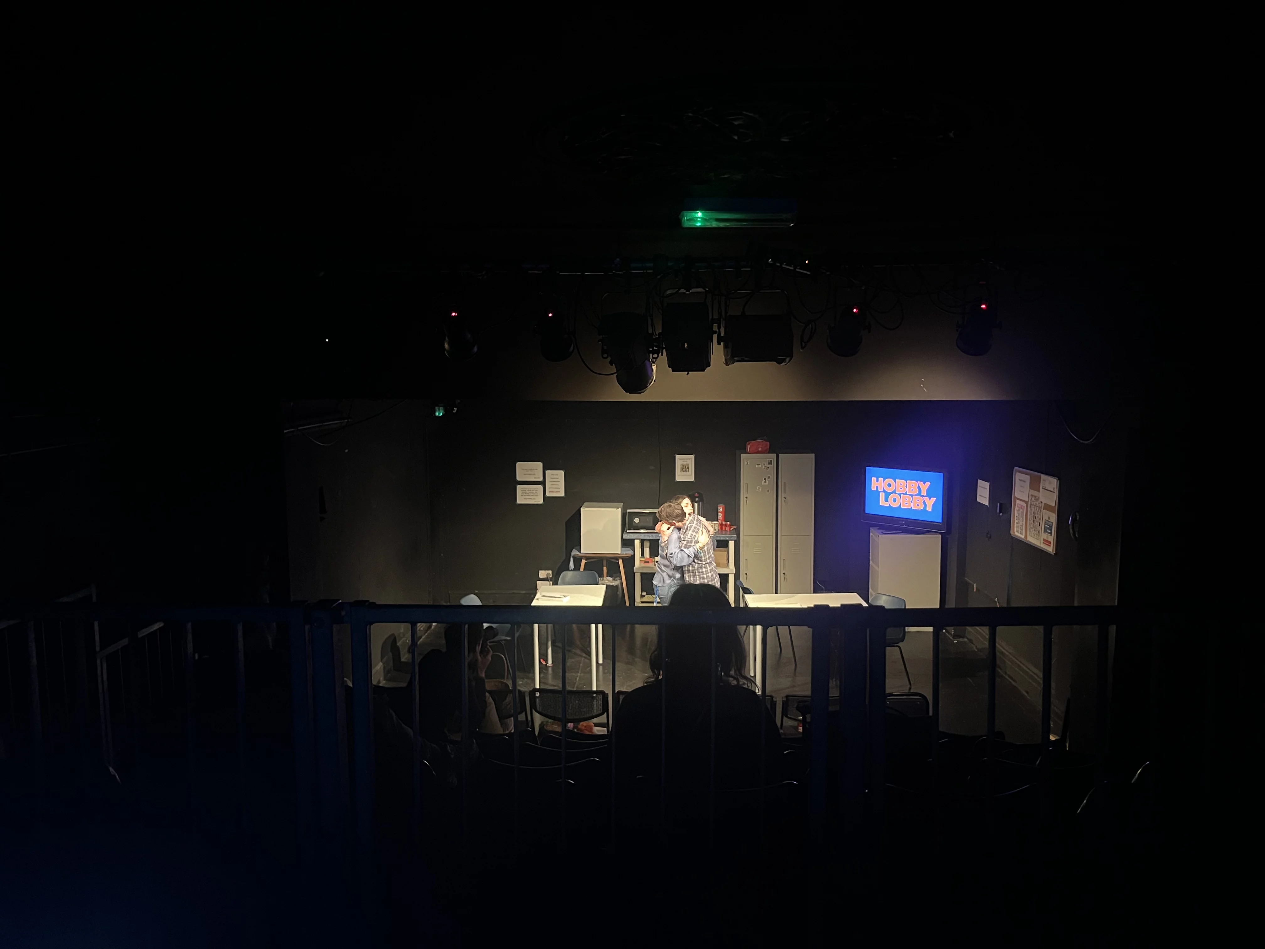 A stage photo from the top of the seats. A man and a woman hug onstage. The stage is set like a staff room, there is a glowing TV displaying a Hobby Lobby logo.