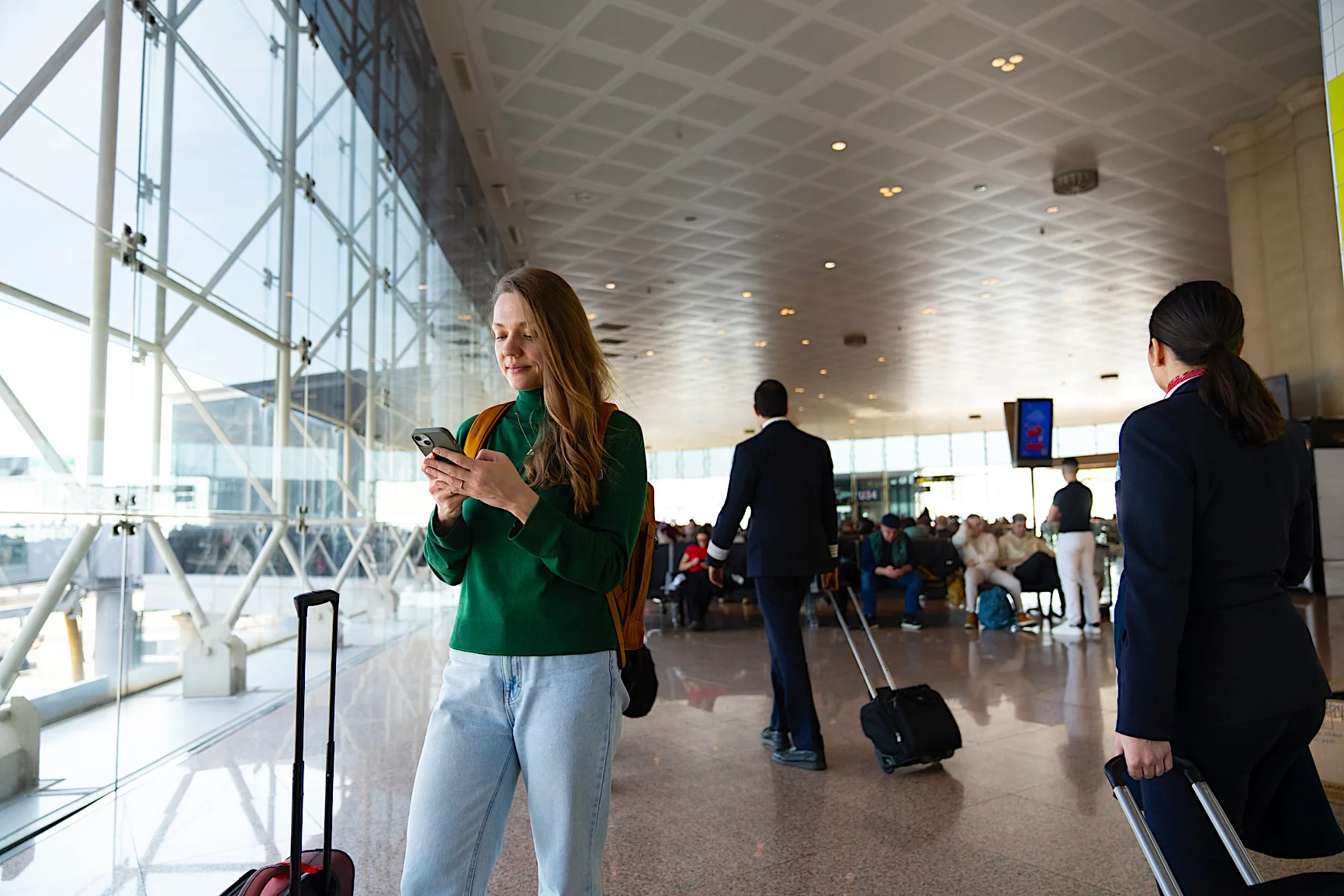 Traveler checking their phone inside a modern airport terminal while waiting with luggage.