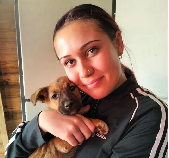Adoption three: Dog. A young lady with a brown ponytail has adopted a small brown puppy. She has blue sharp eyes and sharp eyebrows, a nose piercing and Addidas clothing on. The male puppy is a few weeks old and you can see the young woman will love him for ever!