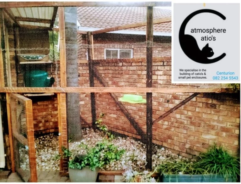 First example of a catio that was built for a client: The catio is small and triangular. It has a rocky floor. The door is to the left and is one meter tall. This door is small and is meant for the animals. There is a small green cat home inside on a ledge within the catio. The catio is surrounded by wire and is made with wooden planks as well.