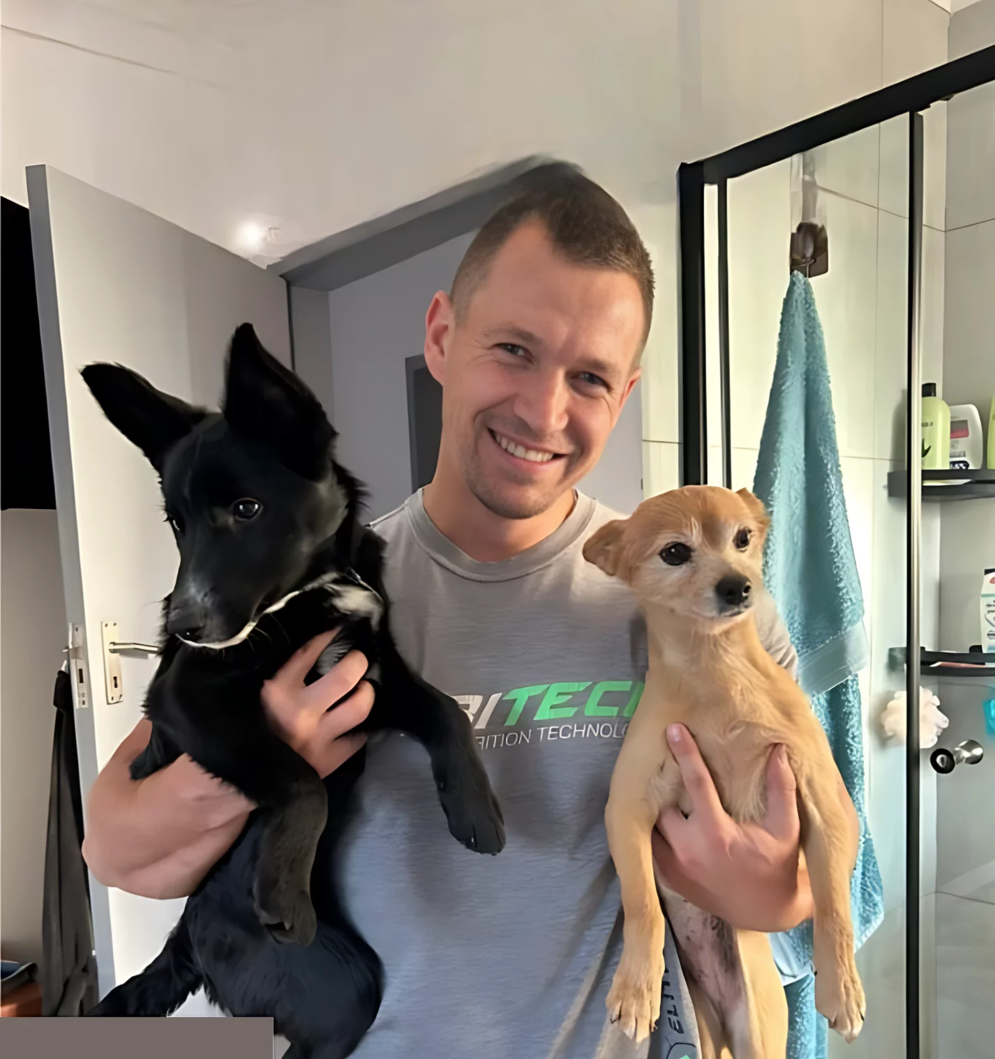 Adoption five: two dogs. A bachelor guy adopted two puppies with different breeds. We do not know the breeds however the one that he is holding in his left side is a baby pitch black puppy with sharp big ears that looks very fit and the other one that he is holding to his right side is a very light brown doggy that looks like a puppy Jack Russel. The man took the picture at home and is smiling happily about his new buddies!