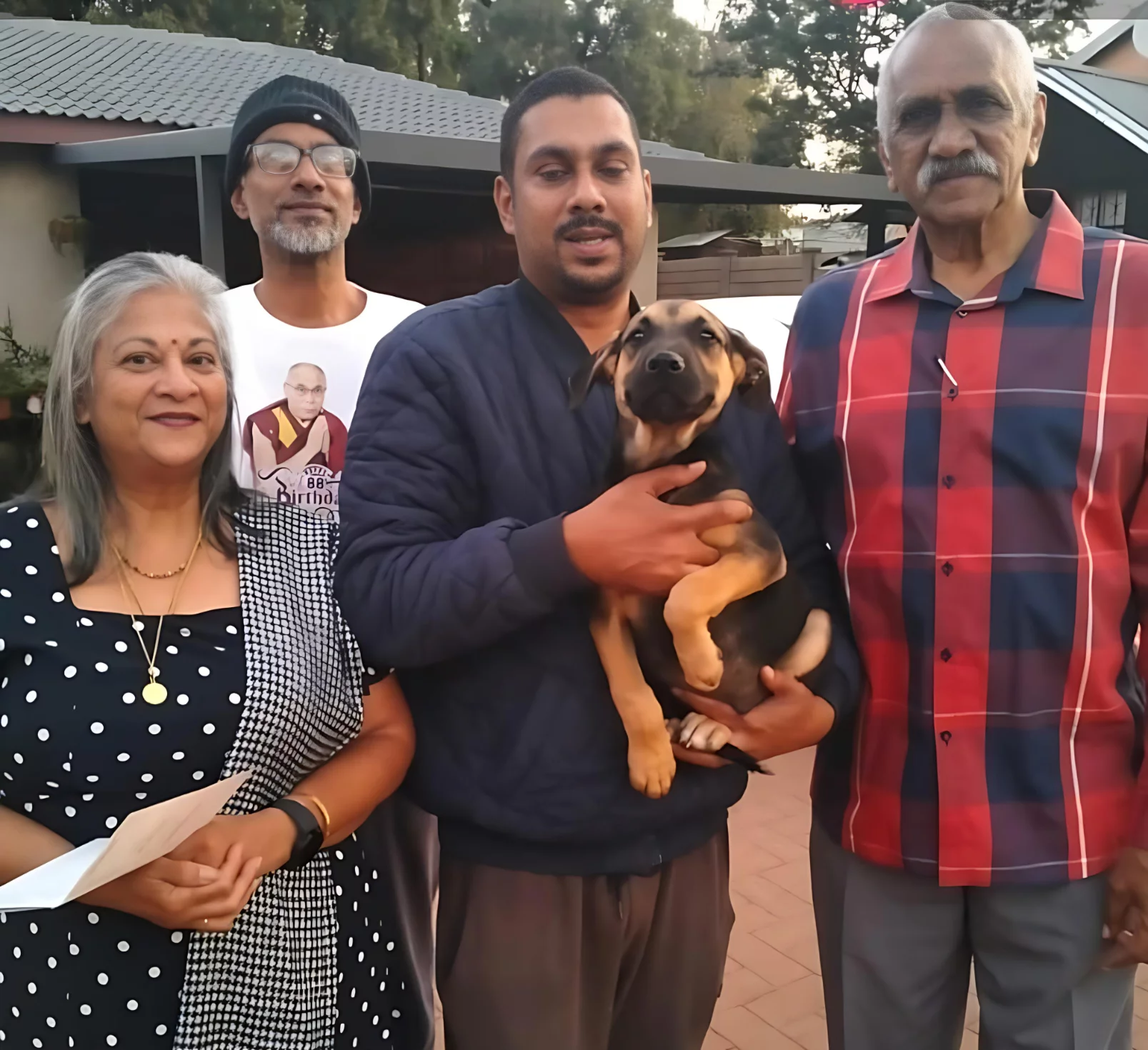 Adoption eleven: Dog. A family of four adopted a small black and brown puppy. The family consists out of an older happily married couple and their two adult son's. The puppy is also smiling into the camera along with is new family.