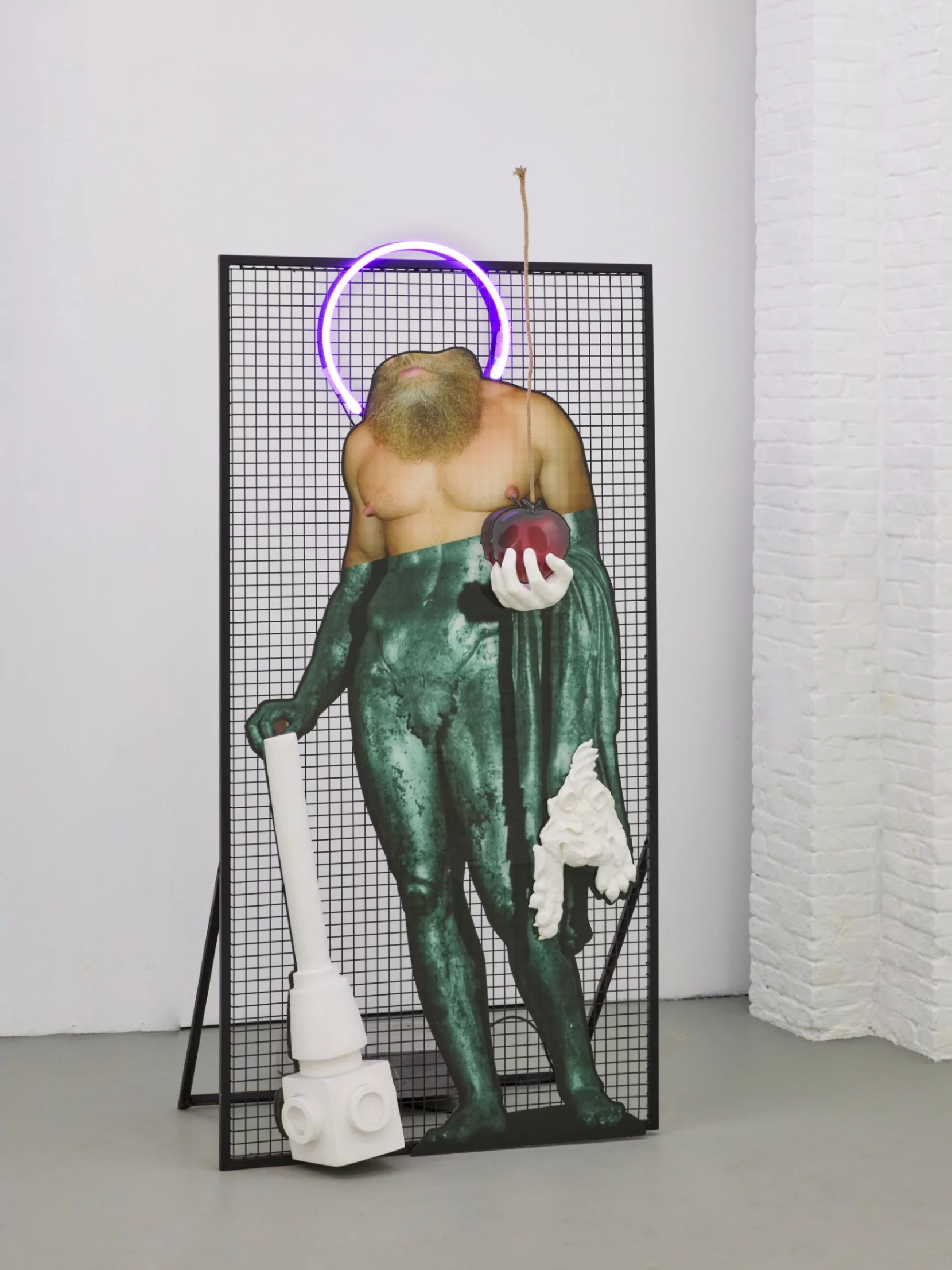 “Byzantine Conundrum,” 2024, a mixed-media sculpture by emerging contemporary sculptor Grigorios Ilaridis at Base-Alpha Gallery, Antwerp. This collage-based sculpture features a 2D headless figure inspired by the Greek hero Heracles, positioned against a black metal grid. A neon purple halo encircles the missing head, inspired by Byzantine iconography. Three prominent 3D elements protrude from the figure: a white plaster club, which the figure seemingly leans on in contraposto, in the right hand, a plaster left hand gripping a 2D red acrylic "poisoned apple" that gives the illusion to be hanging from a string that originates mid-air above the sculpture, and a plaster lion’s head, protruding from the lionskin draped around the arm holding the apple. The upper part of the figure transitions from human into green bronze legs, adding texture and depth, while the white protruding elements contrast dynamically with the bright 2D collage base. Dimensions: 205 x 95 x 60 cm.