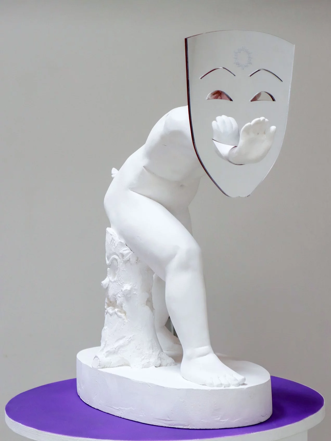 "Double Act: Jokers and Sobbers," 2023 – A sculpture by emerging contemporary sculptor Grigorios Ilaridis showcased at Art Brussels 2024 with Base-Alpha Gallery. This artwork features a white plaster putti figure in a bent-over pose, extending its arm to hold a two-dimensional mirrorplexi Greek theatre mask. The hand of the figure pierces through the mask's mouth, reaching toward the viewer, creating a dynamic interplay of classical motifs and contemporary reflection. Materials: Acrylic, plaster. Dimensions: 55 x 24 x 42 cm.