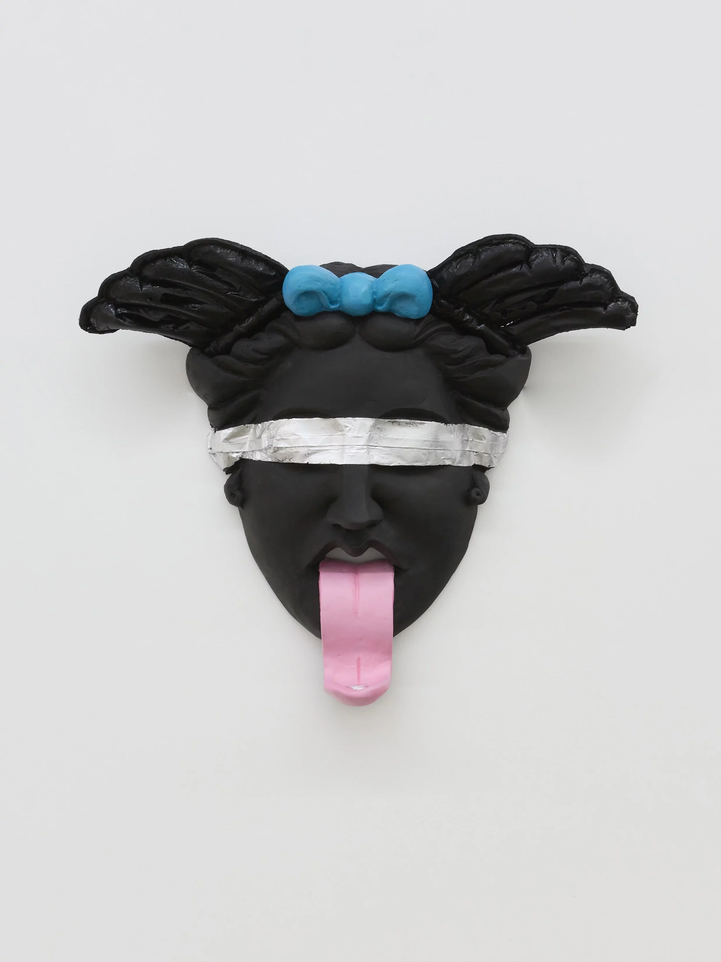 "Oral Tradition II," a matte black mask sculpture by emerging antwerp artist Grigorios Ilaridis at Base-Alpha Gallery in Antwerp. This mask has a Medusa-like quality, featuring a protruding pink tongue, a turquoise bow, and shiny black polyurethane wings framing the hairline. A silver tape band covers the eyes, and a small silver coin depicting Homer sits on the tongue. Materials include acrylic, resin, polyurethane, and silver leaf. Dimensions: 55 x 65 x 13 cm.