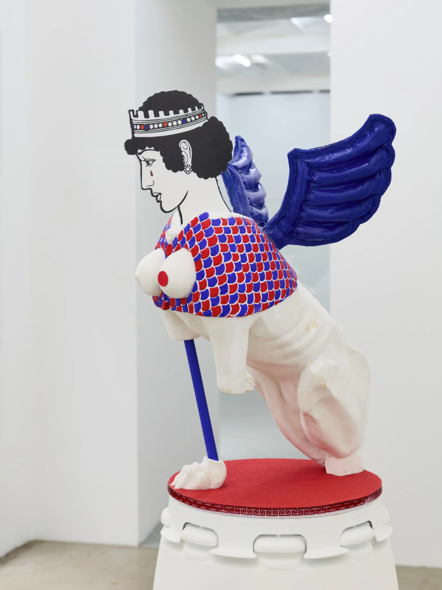 "Rub Me the Right Way," sculpture by Grigorios Ilaridis, exhibited at Base-Alpha Gallery in Antwerp. This acrylic resin sphinx figure features a missing right front paw, with the left paw replaced by an ultramarine blue metal rod ending in a clawed sphinx paw. The armor is crafted with red and blue scale patterns, and two white breasts protrude on the chest, one adorned with a red coin depicting Homer’s head. A tail wraps around the left hind leg, and balloon-like polyurethane wings extend from the spine. The 2D polycarbonate head shows a crowned female figure with a blood-red tear. Dimensions: 172 x 75 x 82 cm.