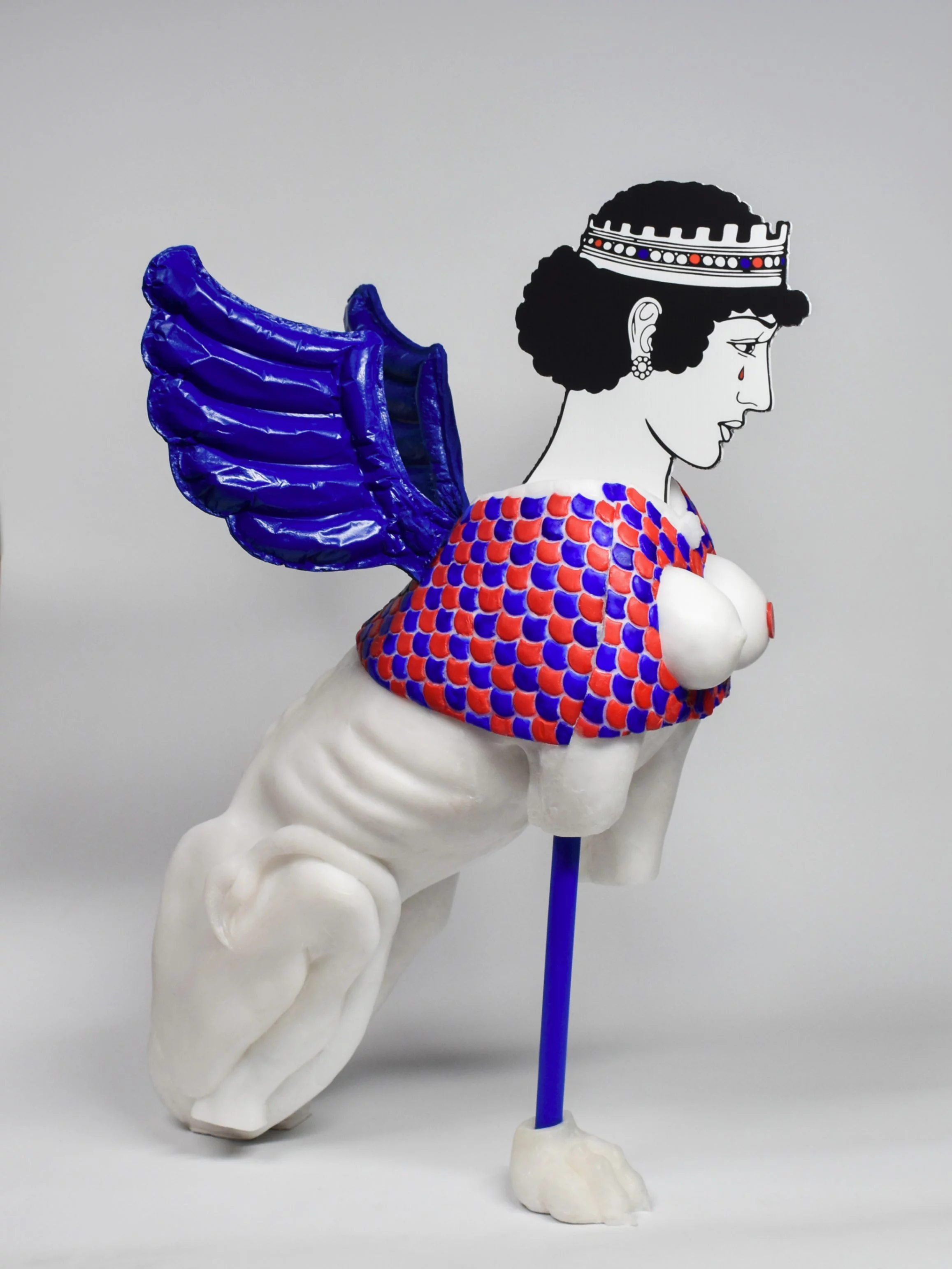 "Rub Me the Right Way," sculpture by contemporary sculptor Grigorios Ilaridis, exhibited at de Warande, Turnhout. This multimedia sphinx figure features a missing right front paw, with the left paw replaced by an ultramarine blue metal rod ending in a clawed sphinx paw. The armor is crafted with red and blue scale patterns, and two white breasts protrude on the chest, one adorned with a red coin depicting Homer’s head. A tail wraps around the left hind leg, and blue balloon-like polyurethane wings extend from the spine. The 2D polycarbonate head shows a crowned female figure with a blood-red tear. Dimensions: 172 x 75 x 82 cm.
