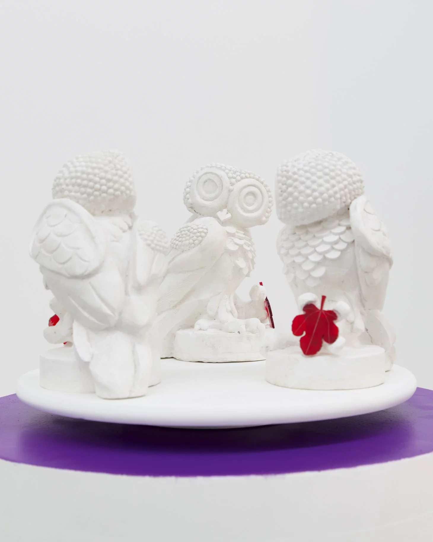"Wisdom in Orbit,” 2024, a sculptural artwork by contemporary artist Grigorios Ilaridis, displayed at Base-Alpha Gallery, Antwerp. This piece features three white resin owls arranged in a circular formation on a purple wooden base, each owl gazing inward and adorned with a mirrored acrylic star on its forehead. The owls hold a vibrant red acrylic fig leaf between them, symbolizing wisdom and unity. Dimensions: 145 x 50 x 50 cm.