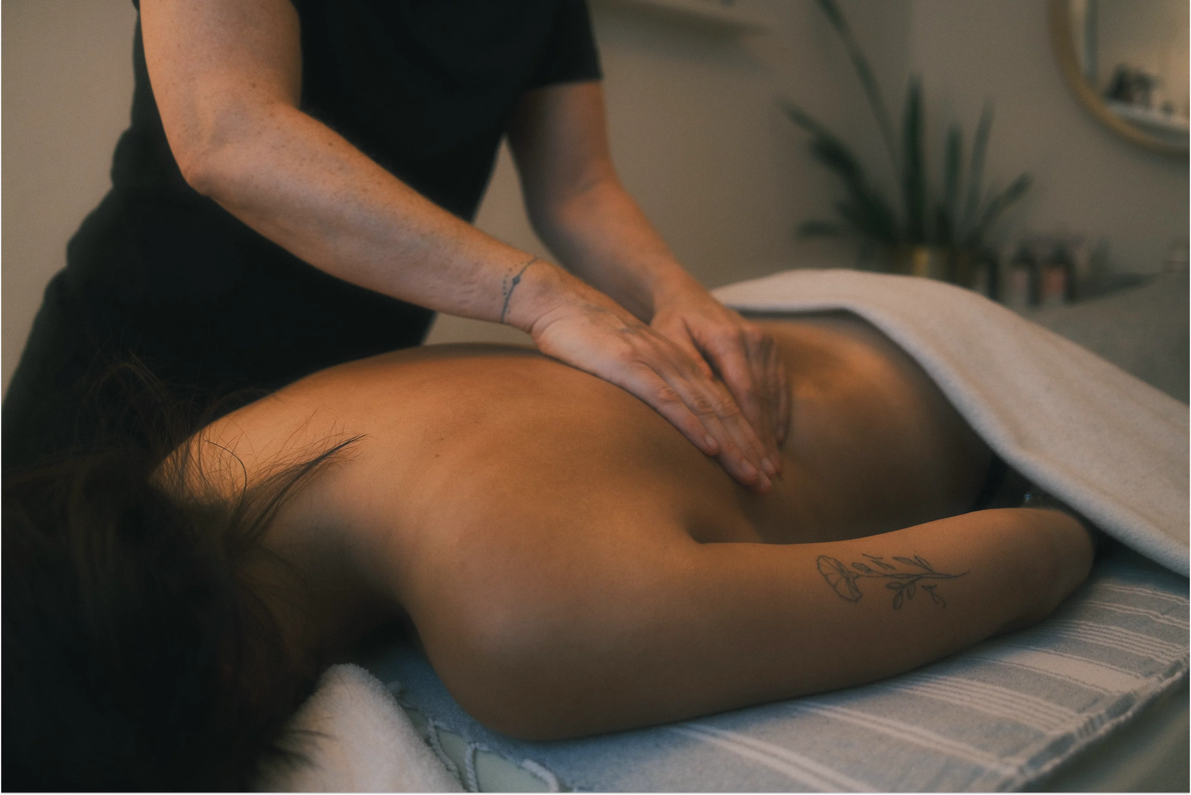 Eine Person liegt mit nacktem Rücken auf einer Massageliege, während Sabine ihr mit beiden Händen den Rücken massiert. Die empfangende Person ist teilweise mit einer Decke bedeckt. Der Raum wirkt warm&nbsp;und&nbsp;beruhigend