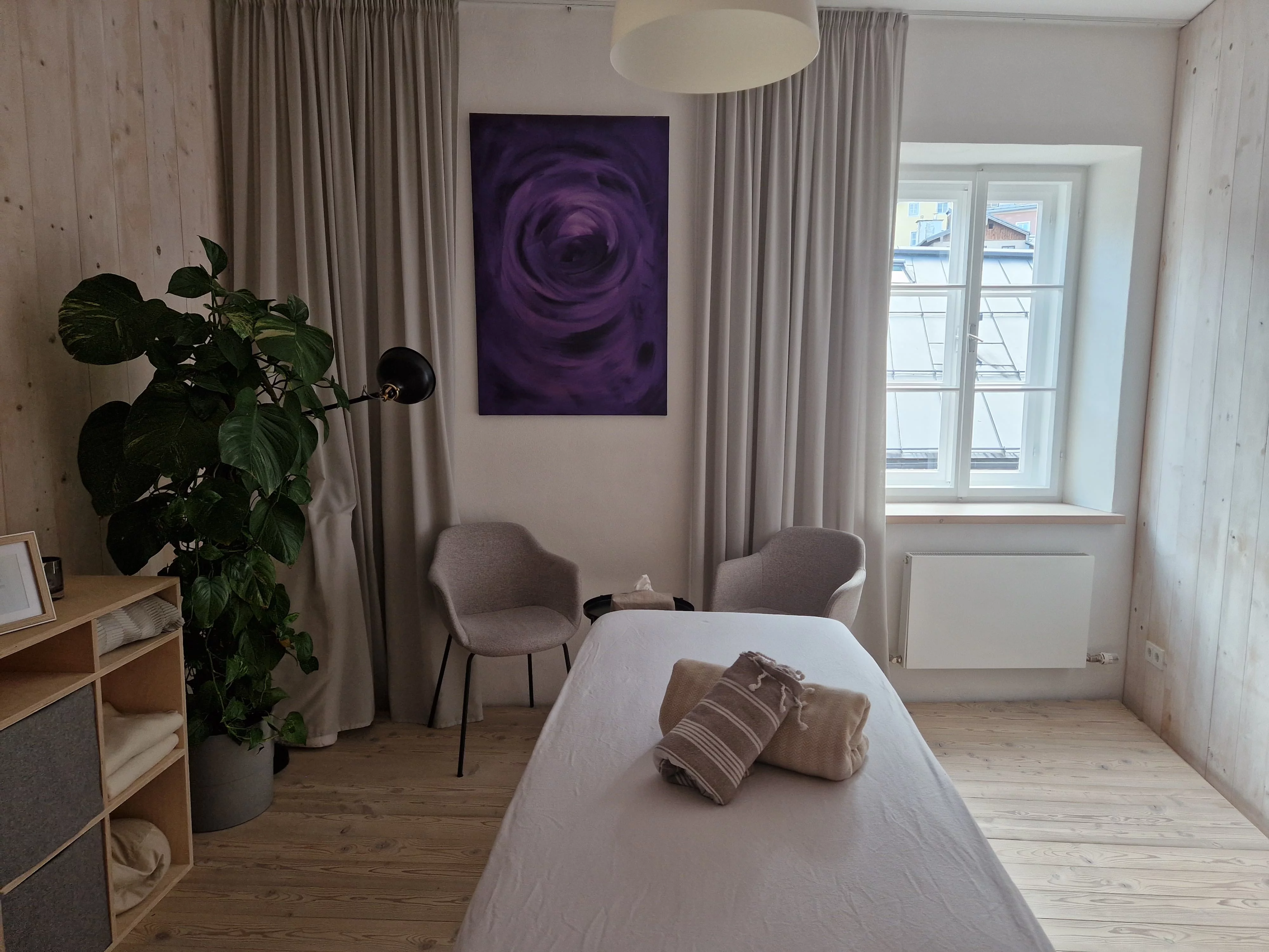 Eine Massageliege in einem gemütlichen, hellen Raum mit Holzboden und umrahmt von einer Pflanze und einer Sitzgruppe.