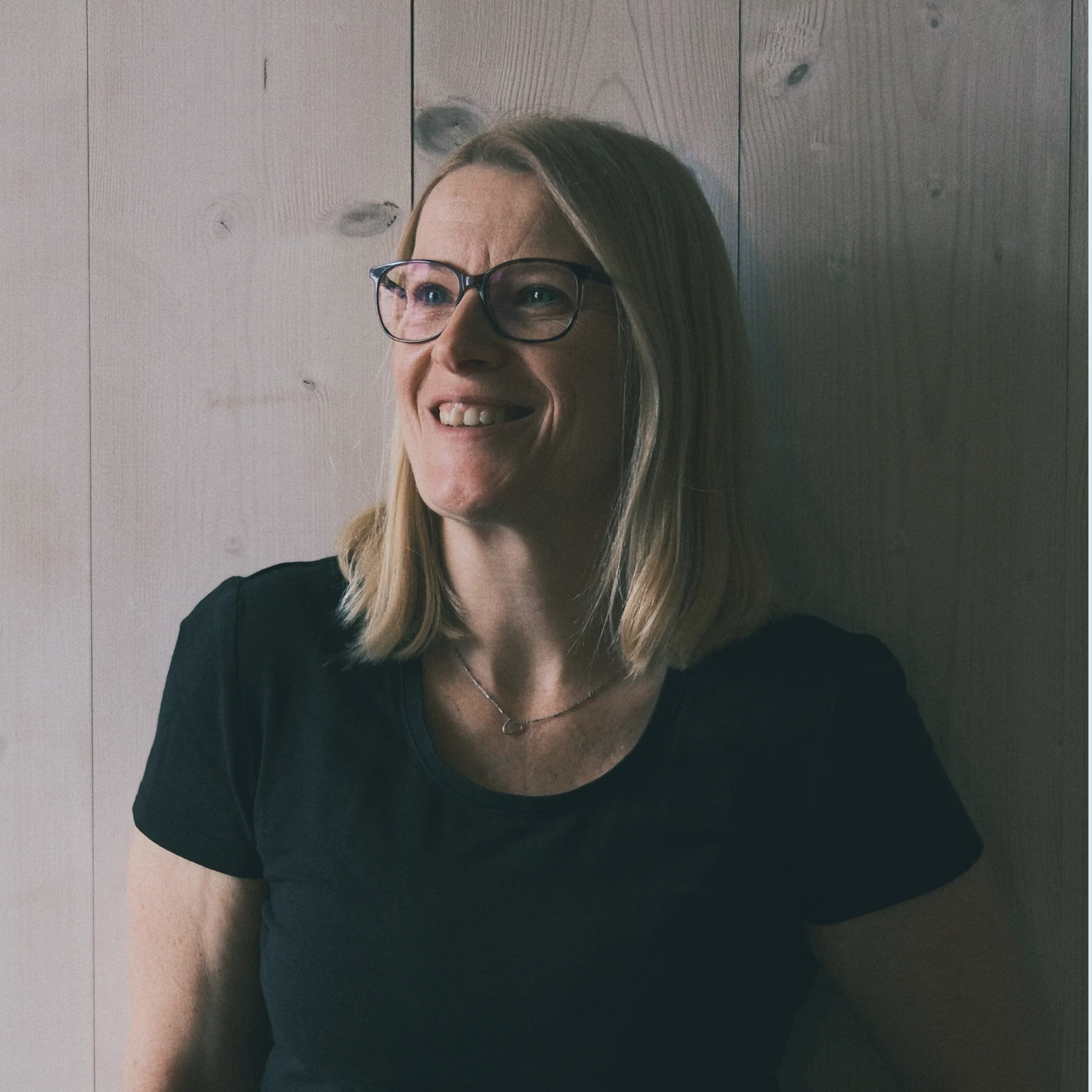 Profilbild von Sabine. Eine Frau mit blondem Haar und Brille steht lächelnd vor einer Holzwand. Sie trägt ein schwarzes T-Shirt und strahlt Ruhe und Zufriedenheit aus.